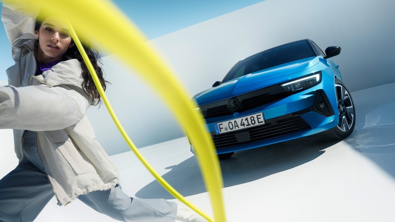 Pogled od spredaj na modro Opel Astro Electric z žensko, ki vrti rumeni polnilni kabel