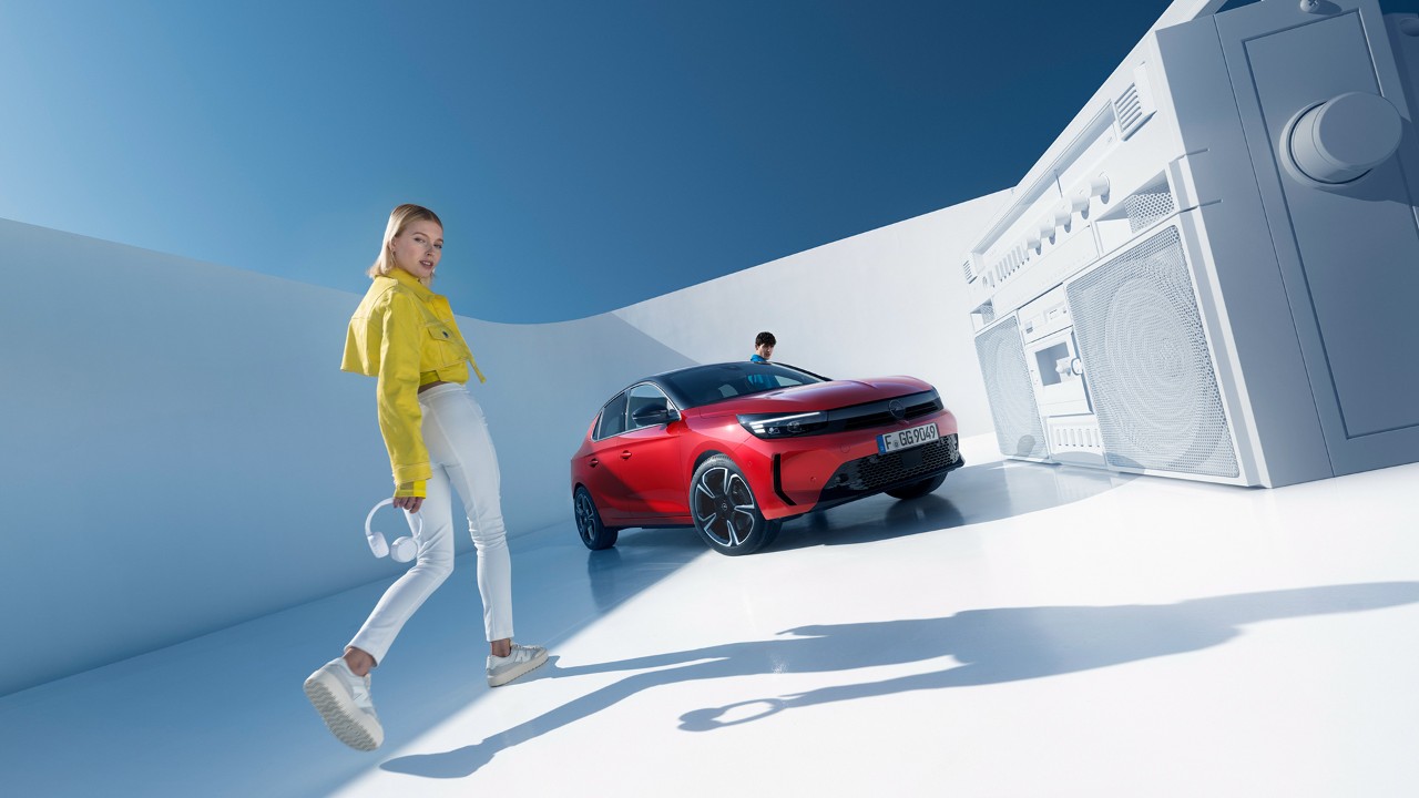 Rdeča nova Opel Corsa parkirana v daljavi z ženskami v rumenih jopičih, ki hodijo proti njej in gledajo v kamero